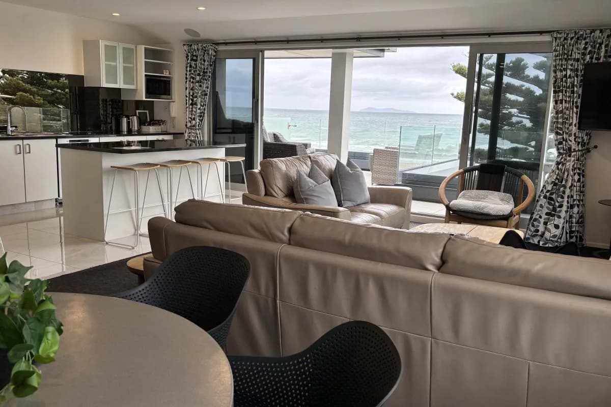 Living area with leather sofas, kitchen island, and ocean view through glass doors.