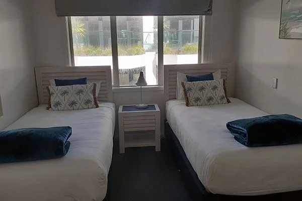 Twin beds with palm-print pillows and teal throws beside a window.