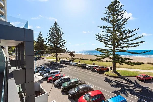 Beachfront view from Pavilion apartments overlooking parked cars, pine trees, and Mount Maunganui beach.